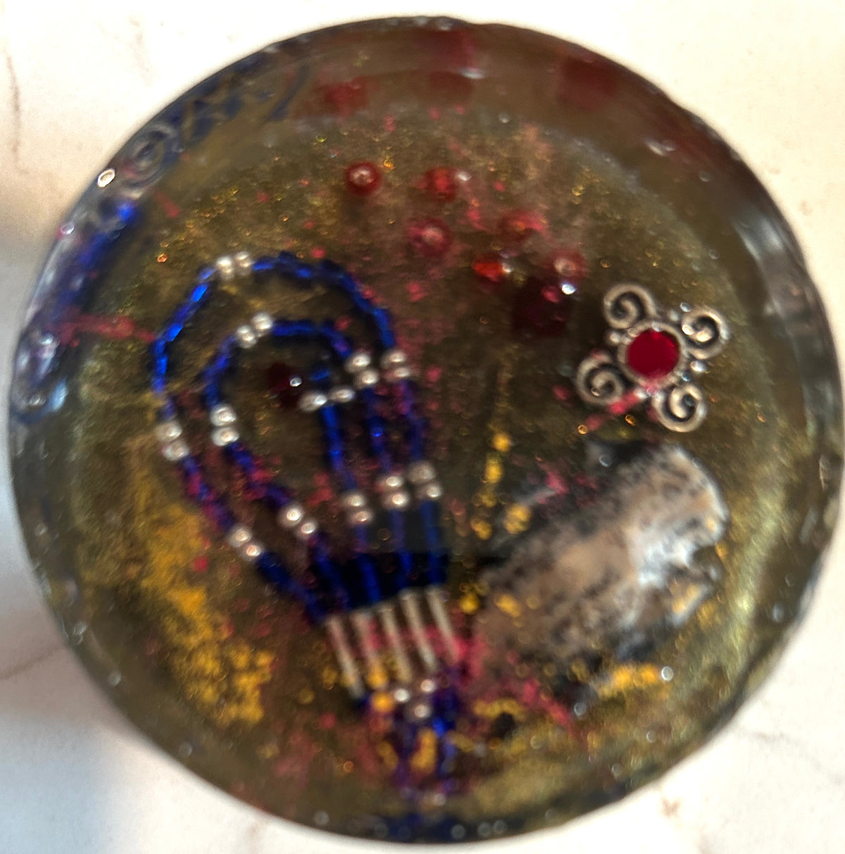 Round resin paperweight with blue beaded heart, silver cross, red crystal filigree flower charm, and gold glitter field