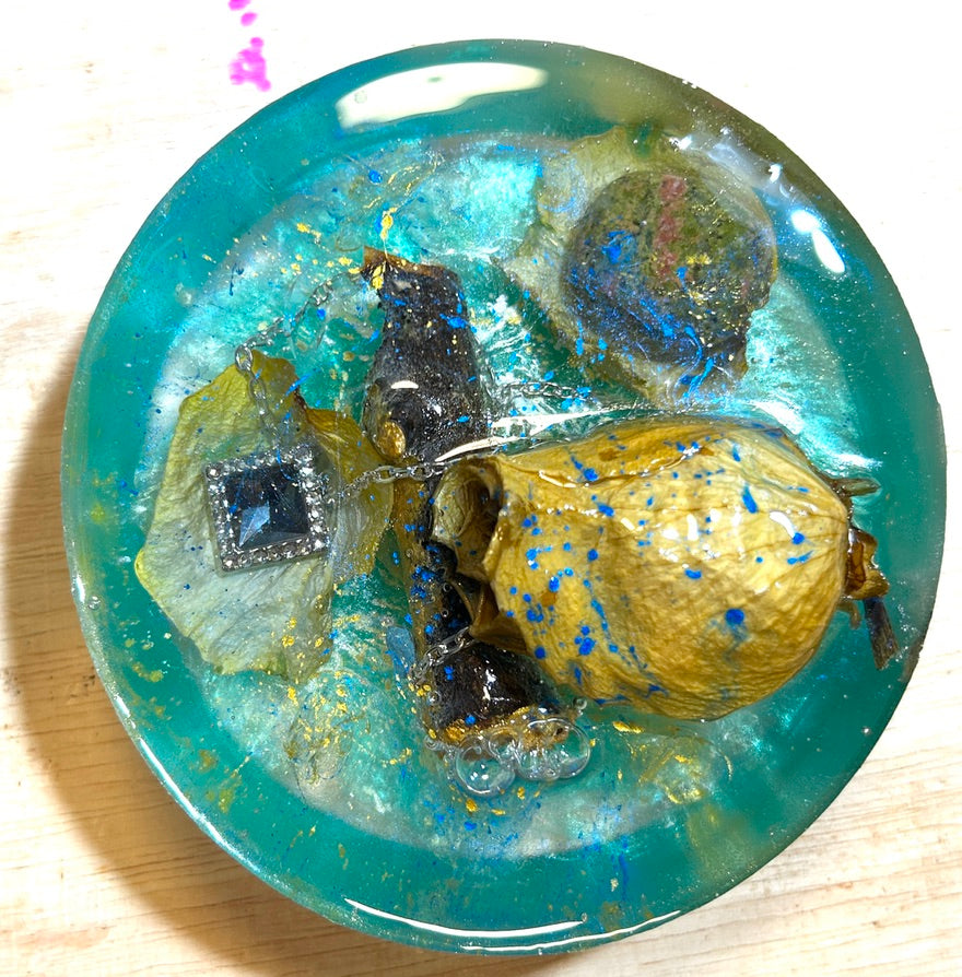 Tidal Rose resin paperweight — overhead view showing resinized golden rose bud with blue glitter, square blue rhinestone pendant on silver chain, dark stone, dried botanicals and bark in vivid teal resin with smooth round edges