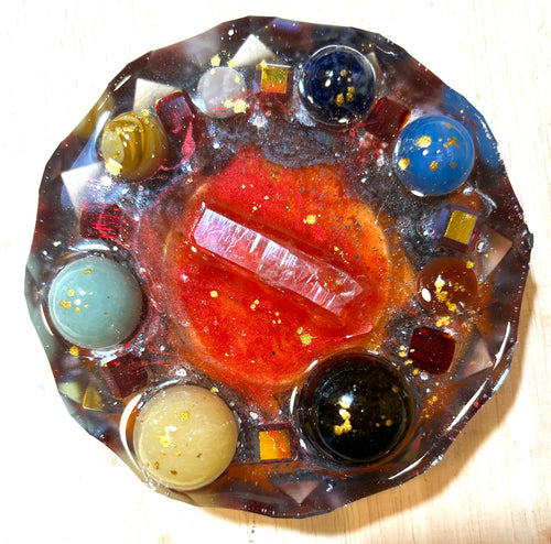 Ember & Obsidian resin paperweight — overhead view showing central selenite crystal point on red shimmer base, surrounded by seven polished stone spheres including blue lapis, aquamarine, black obsidian, and cream, with gold leaf flecks and faceted diamond edges