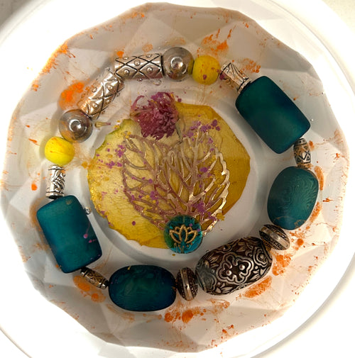 Desert Bloom resin paperweight — overhead view showing resinized yellow leaf with gold filigree tree charm and purple thistle, teal lotus bead, surrounded by ring of teal sea glass, Tibetan silver and bronze beads in clear resin with orange splatter, faceted diamond edges