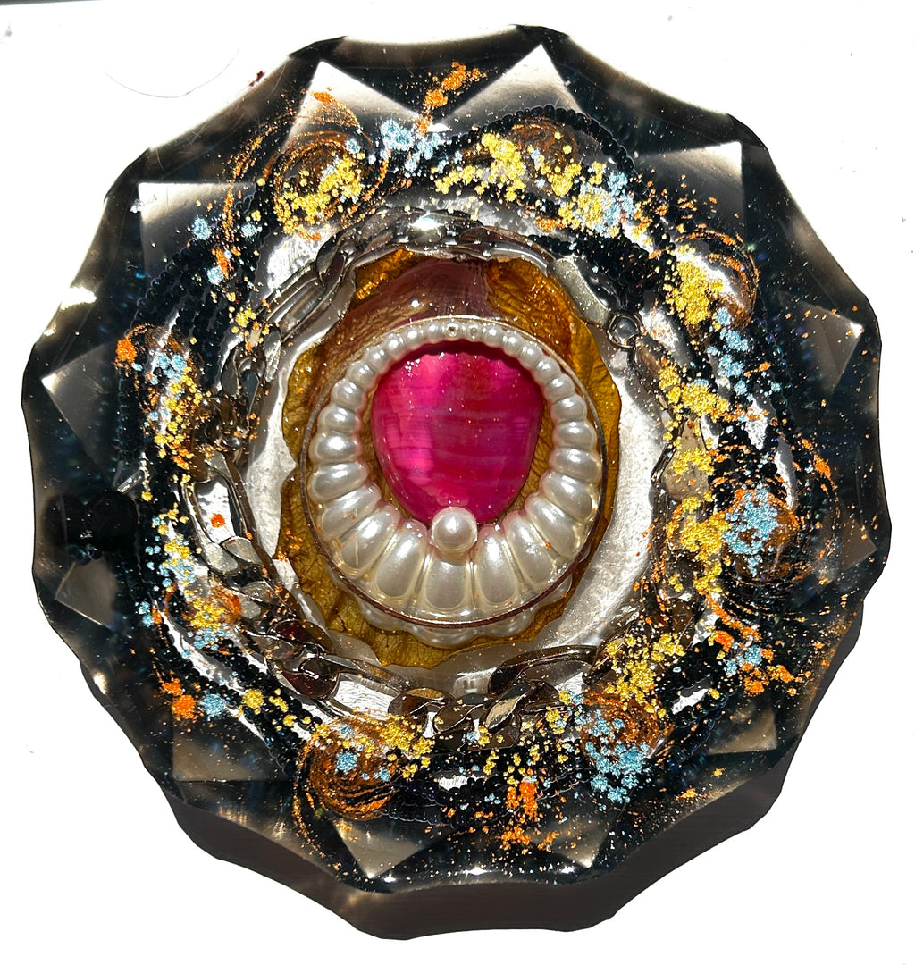 Black Pearl Abyss resin paperweight — overhead view of round disc with faceted scalloped edges showing deep pink magenta teardrop stone in pearl-edged oyster brooch setting with silver serpent chain coiling around it in deep black resin with gold leaf and orange teal glitter