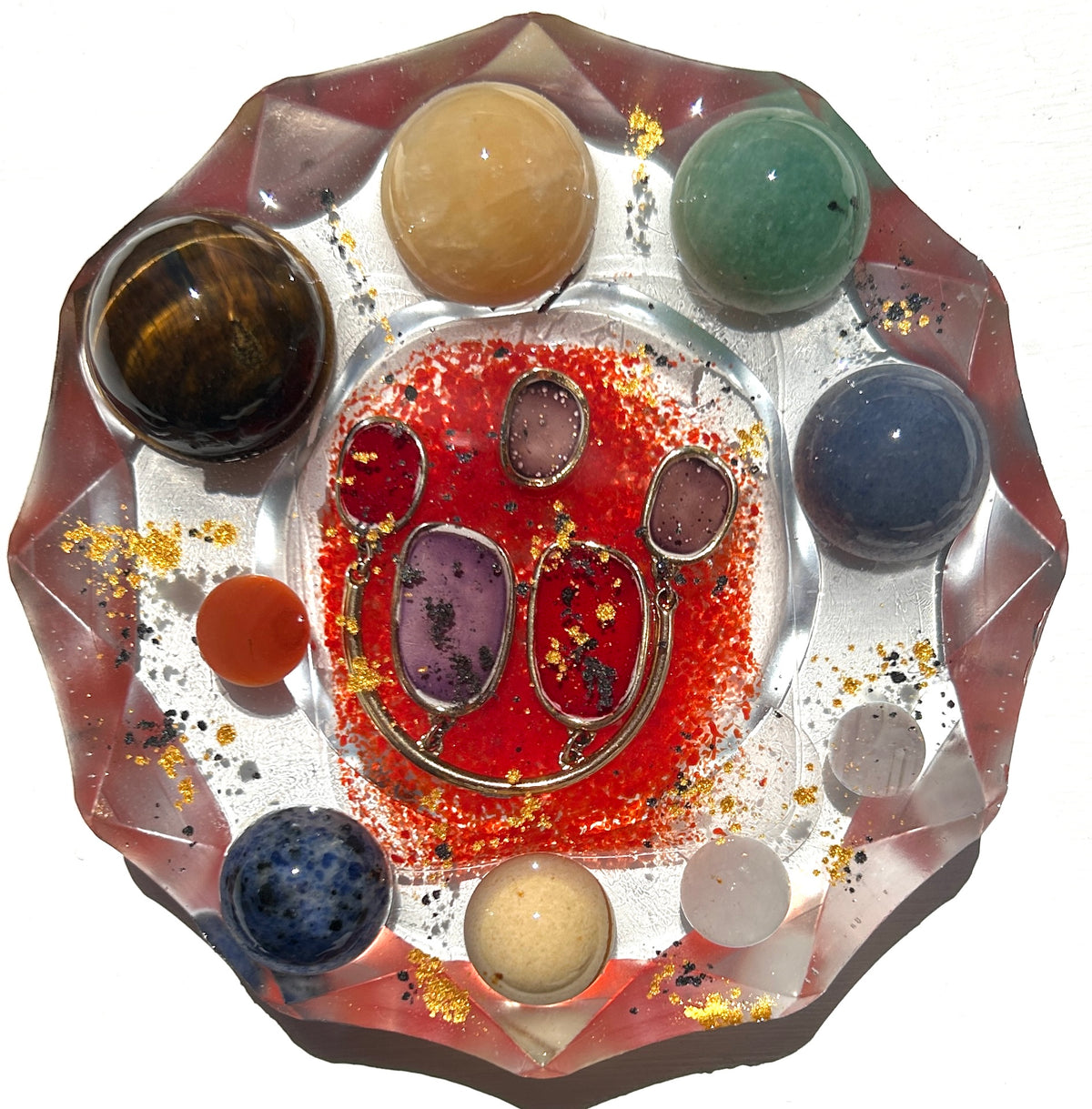 Celestial Orbits resin paperweight — overhead view of large round disc with faceted diamond-cut edges showing 8 polished crystal spheres in a ring surrounding gold-framed amethyst crimson and rose quartz earring set in red crimson glitter resin with gold leaf and black spatter