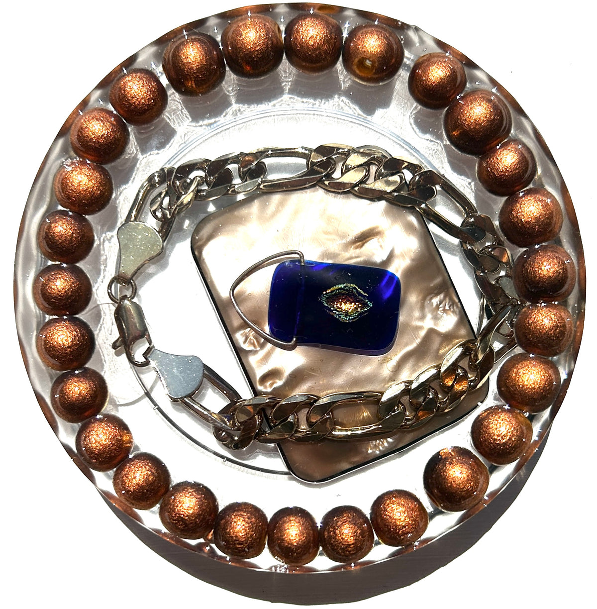 Evil Eye Talisman resin paperweight — overhead view of round disc with smooth edges showing cobalt blue glass evil eye pendant on gold silk square surrounded by heavy silver figaro chain and outer ring of copper bronze metallic shimmer beads in clear luminous resin