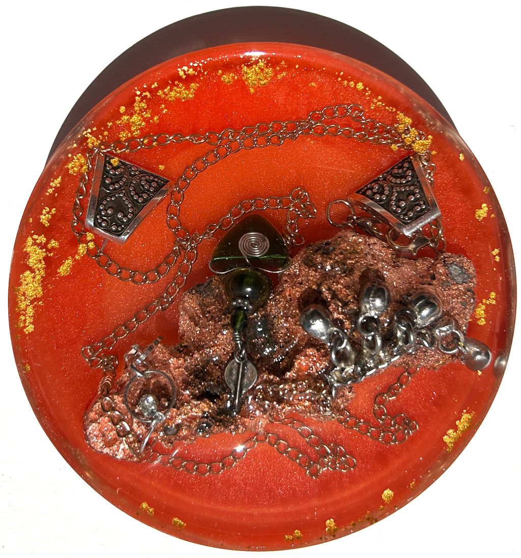 Volcanic Fire resin paperweight — overhead view of round disc with smooth dome edges showing raw dark copper volcanic mineral cluster with wire-wrapped jade pendant, silver mercury glass orbs, two ornate silver filigree triangle pendants on draped silver chain in deep vermillion orange-red resin with gold leaf flakes