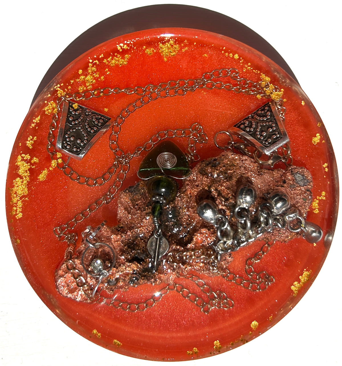 Volcanic Fire resin paperweight — overhead view of round disc with smooth dome edges showing raw dark copper volcanic mineral cluster with wire-wrapped jade pendant, silver mercury glass orbs, two ornate silver filigree triangle pendants on draped silver chain in deep vermillion orange-red resin with gold leaf flakes