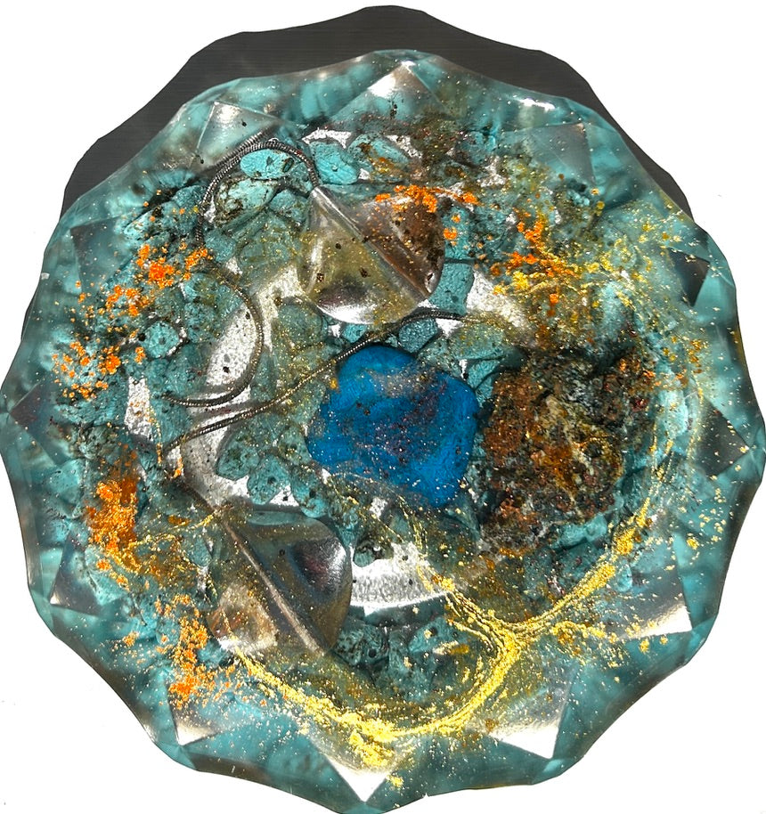 Ocean Geode resin paperweight — overhead view of round disc with faceted scalloped edges and dark charcoal rim showing cobalt blue heart stone, rough turquoise chrysocolla chunks, clear crystal shards, silver rope chain and swirling gold vortex in clear aqua teal resin with orange glitter and gold leaf