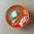 Orange resin paperweight with pale seashells, turquoise-colored flecks, a clear crystal-like point and a pale pink accent.