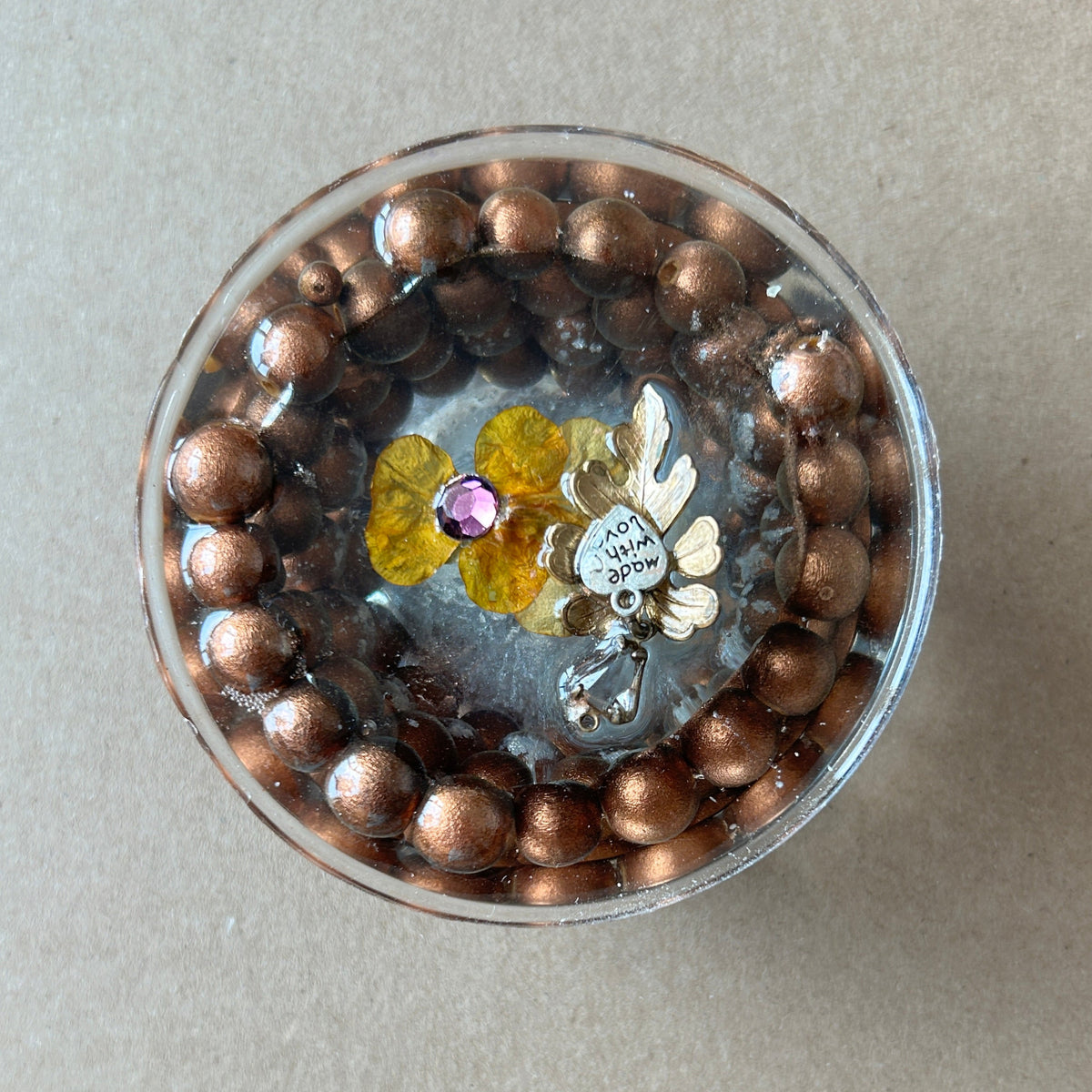 Round paperweight with copper-brown bead border, dark inner ring, yellow flower detail and metallic charms.