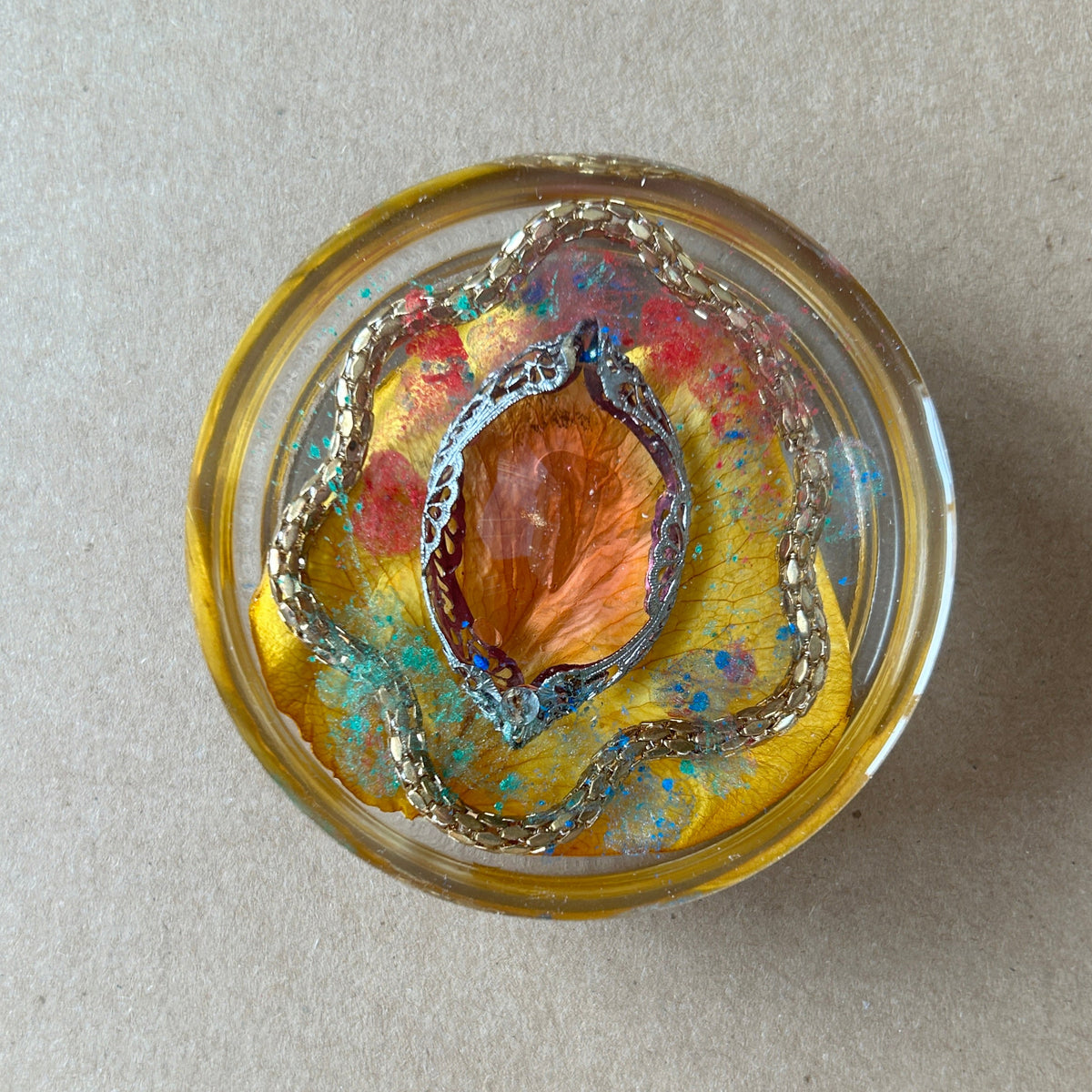 Yellow resin paperweight with orange-gold botanical center, ornate oval silver-toned frame and looping chain.