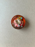 Amber-orange paperweight with gold-toned leaf charm, pink textures and three small colorful floral-patterned accents.