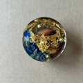 Round paperweight with ornate gold-toned scrollwork, metallic beads, blue fragments and brown wood-like forms.
