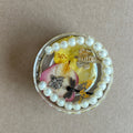 Round paperweight with white bead border, pink and yellow floral details, dark dragonfly, tiny bee and striped shell.