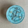 Pale aqua round paperweight with looping silver-toned chain, pearly white accents and two golden knot details.
