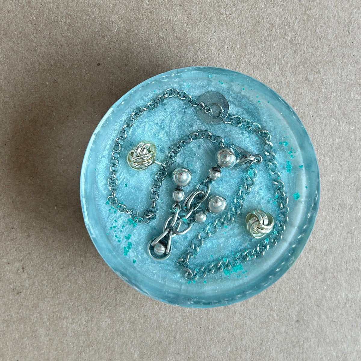 Pale aqua round paperweight with looping silver-toned chain, pearly white accents and two golden knot details.