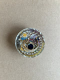 Eye-shaped jeweled ornament with deep blue center and a ring of clear sparkling accents in resin