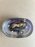 Oval violet-tinted paperweight with a shell, dark beads, pearly accents and an ornate silver-toned charm