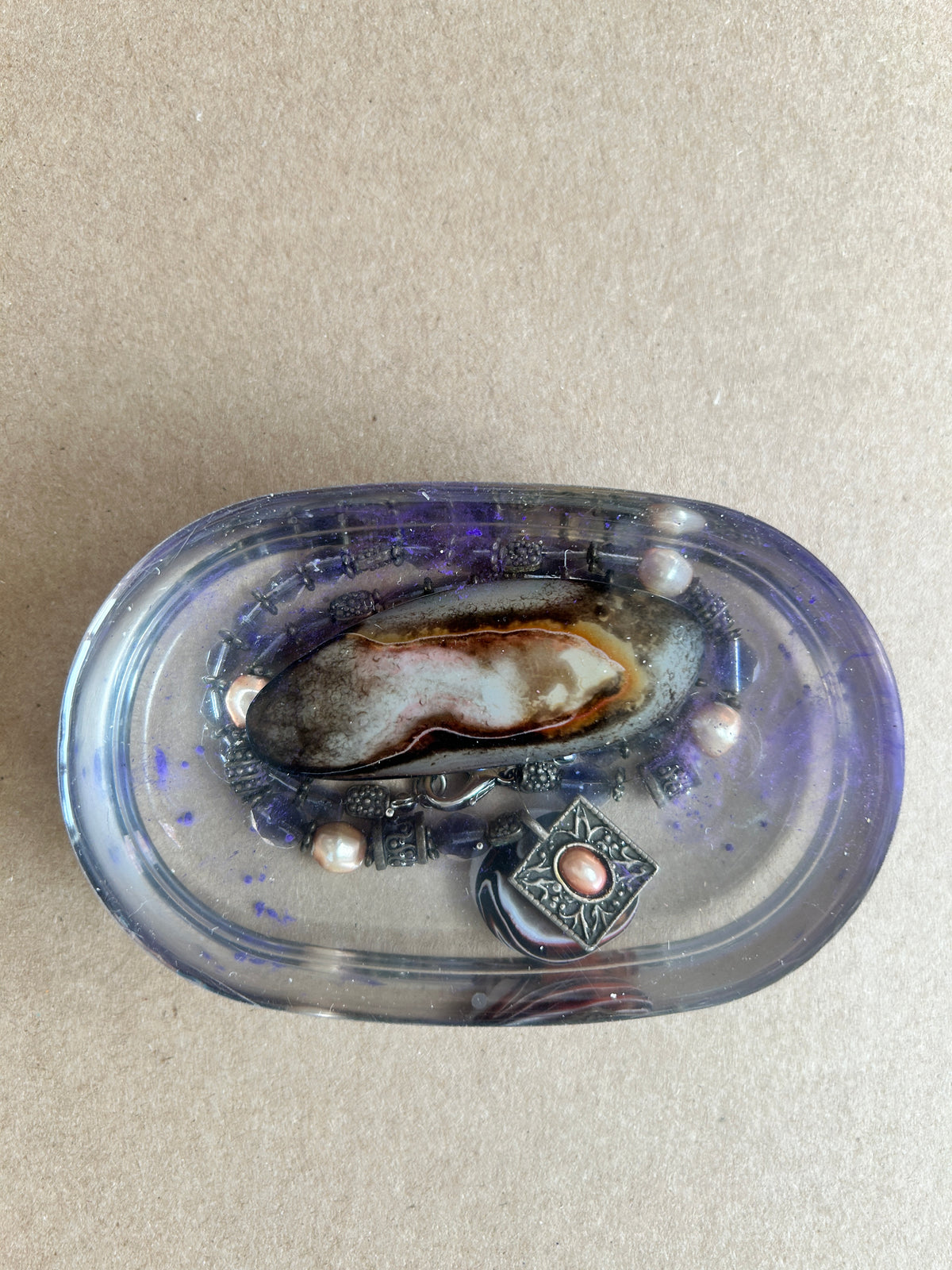 Oval violet-tinted paperweight with a shell, dark beads, pearly accents and an ornate silver-toned charm
