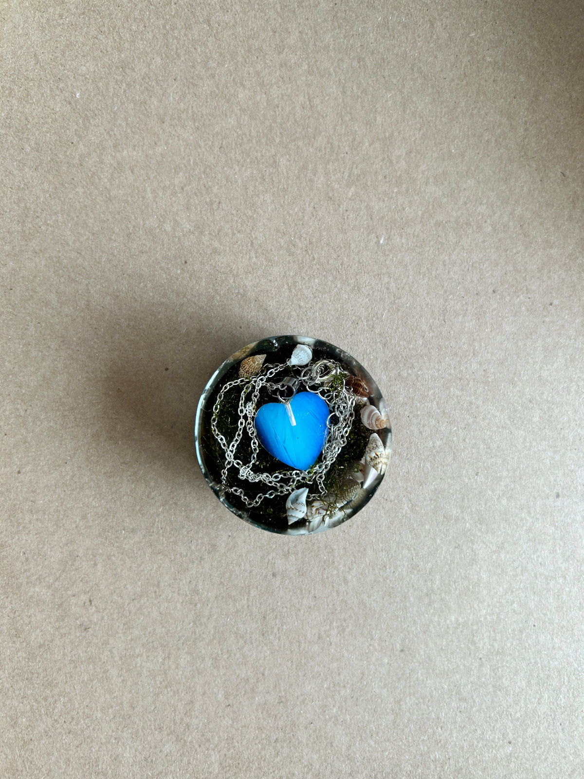 Blue heart surrounded by silver-toned chains and small shells in a dark resin paperweight