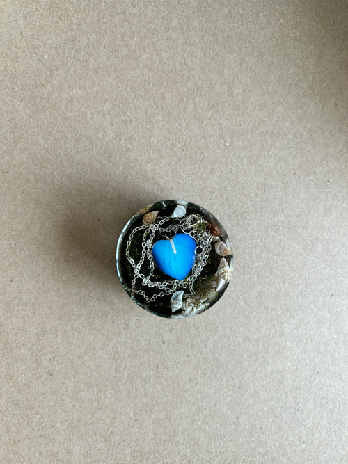 Blue heart surrounded by silver-toned chains and small shells in a dark resin paperweight