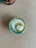 Pearly white fan around a gold-toned oval frame with a luminous center in pale aqua resin