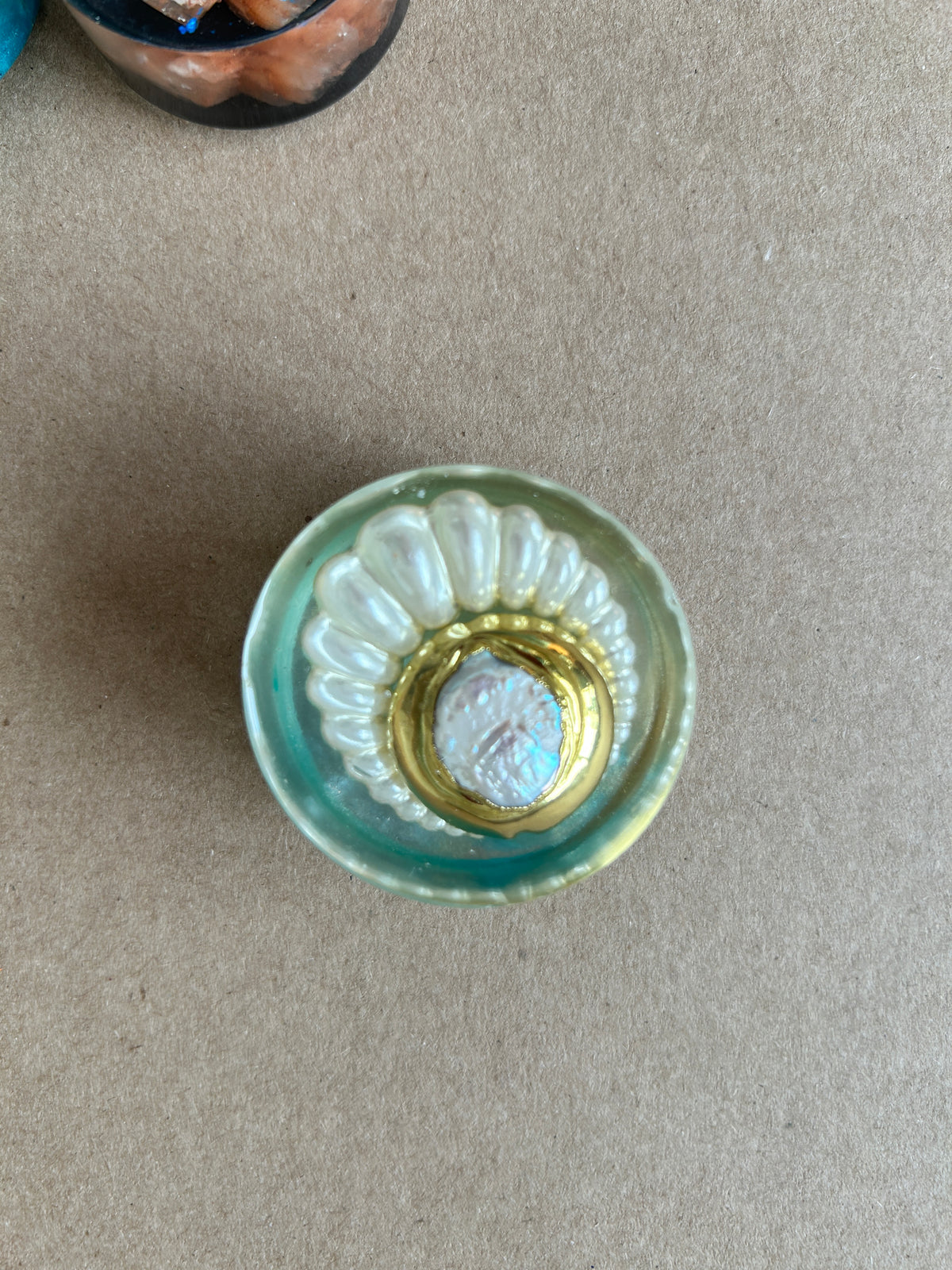 Pearly white fan around a gold-toned oval frame with a luminous center in pale aqua resin