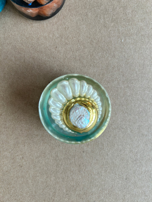 Pearly white fan around a gold-toned oval frame with a luminous center in pale aqua resin