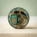 Teal Treasury paperweight with ornate teal centerpiece, gold-toned chain, tiny clear bottle and golden flecks on a light background